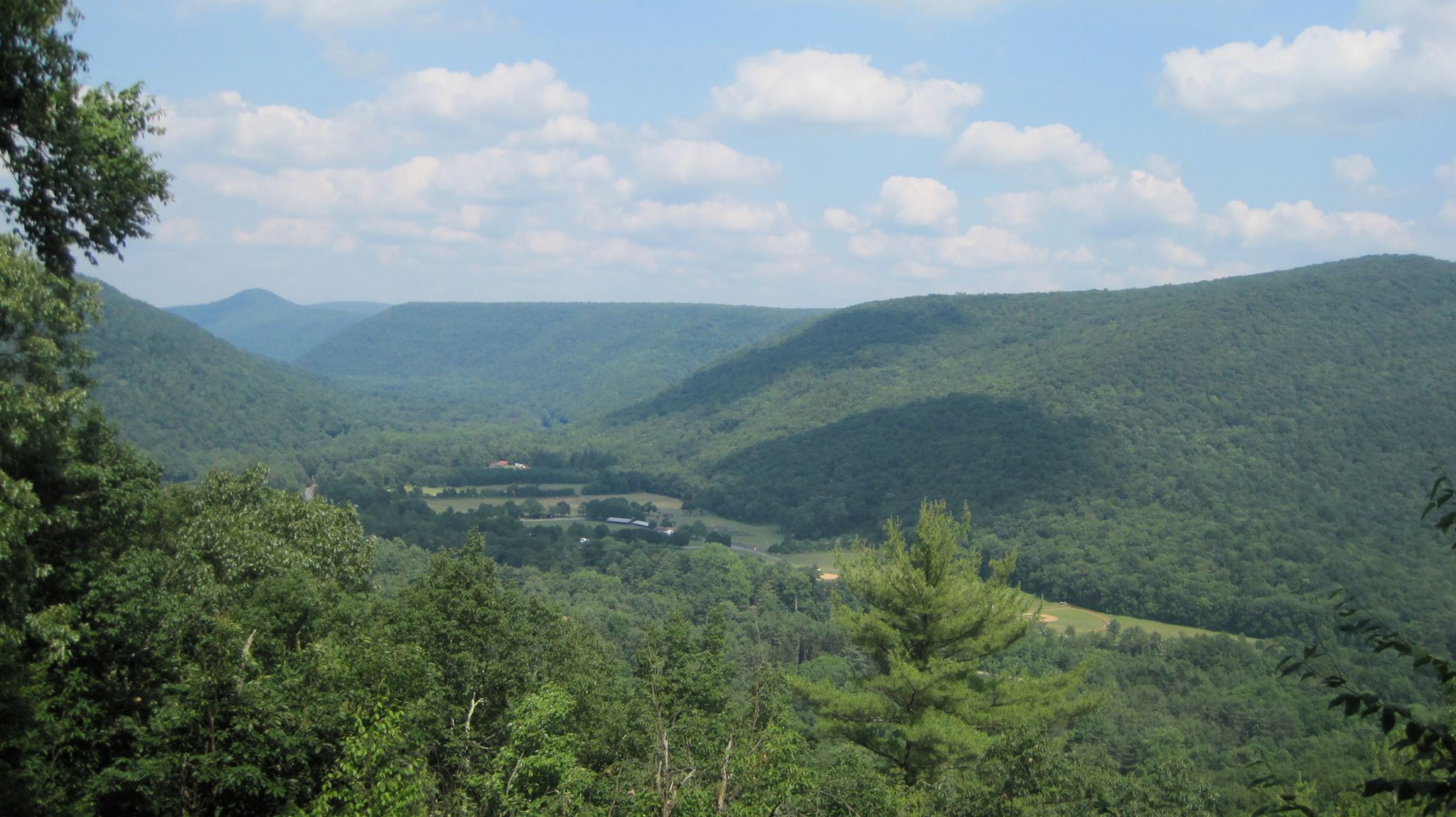 Photograph of the Loyalsock Valley in Lycoming County, PA from Doe Pen Vista. Appx. 2000' tall rounded mountains in full summer foliage surround a farm valley cut by the Loyalsock Creek.